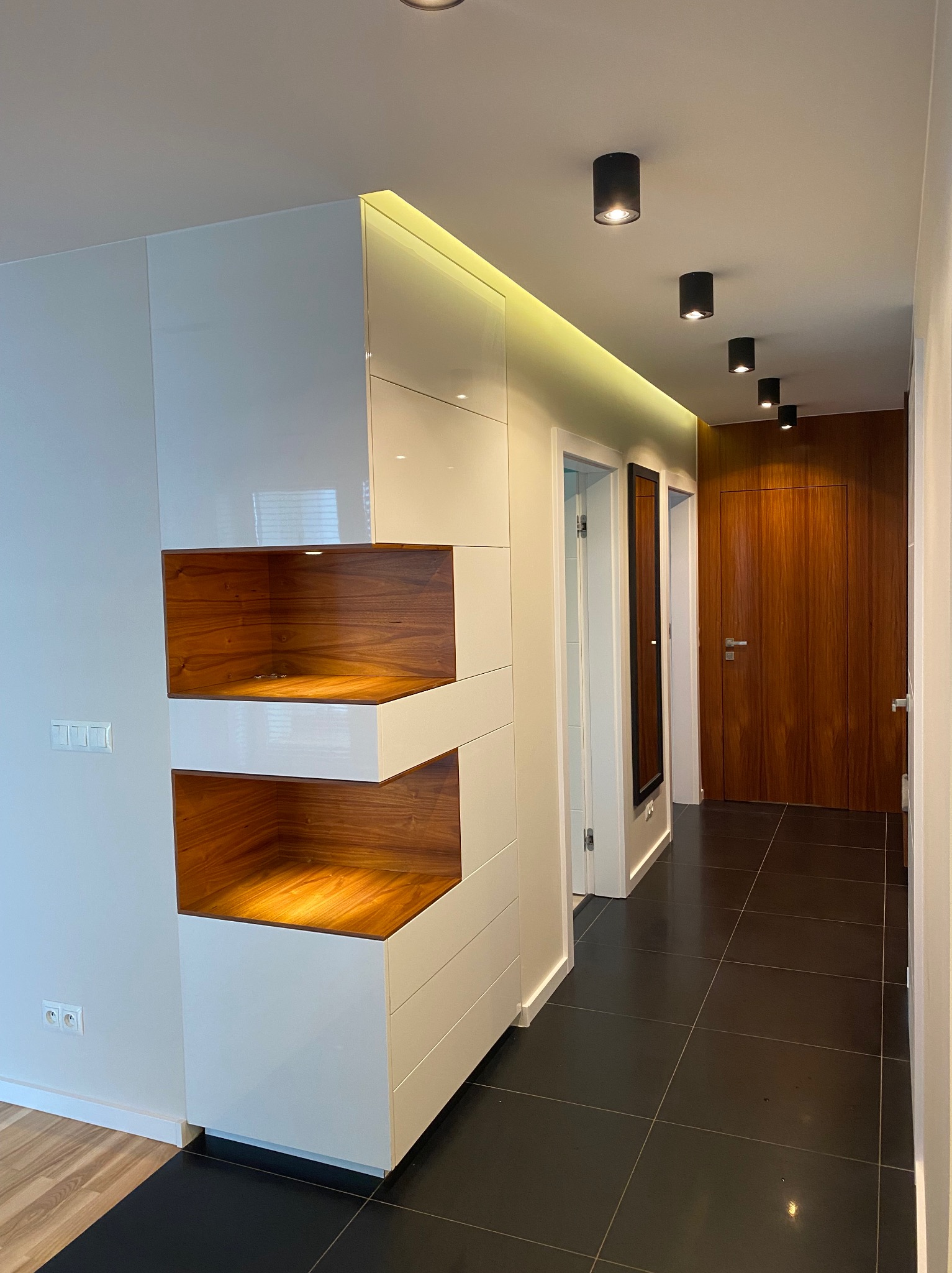 Nowoczesny przedpokój z zabudową meblową: białe szafy z drewnianymi wnękami, czarna podłoga, oświetlenie punktowe. Minimalistyczny design, eleganckie wykończenie.