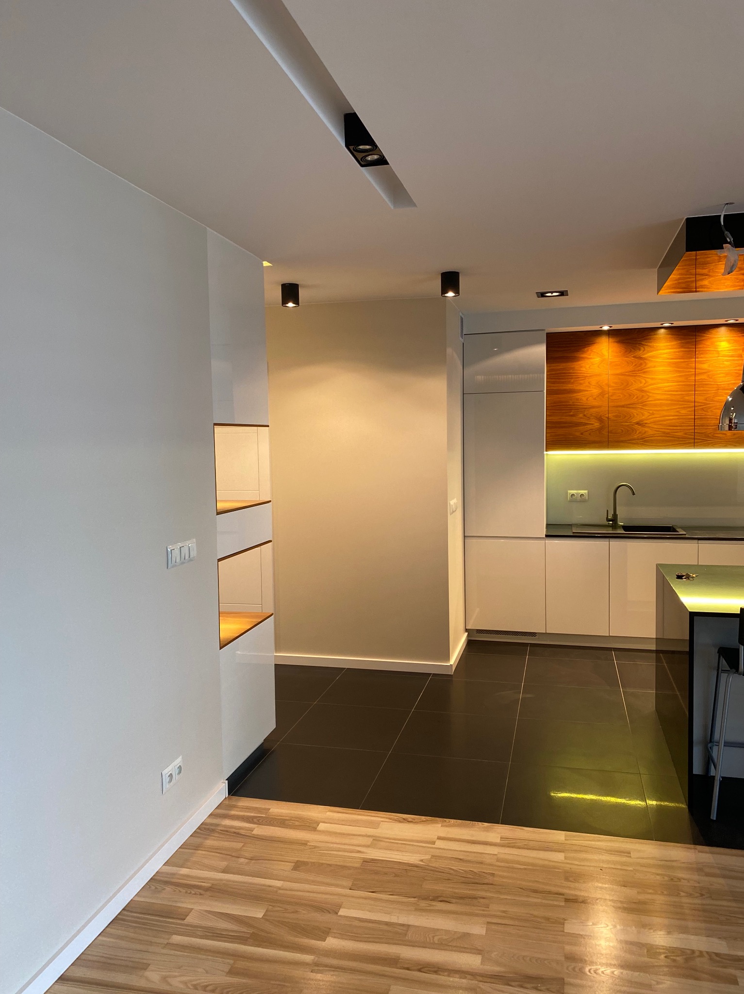 Nowoczesna kuchnia z drewnianymi akcentami i czarną podłogą w Warszawie. Białe szafki, oświetlenie LED i minimalistyczny design.