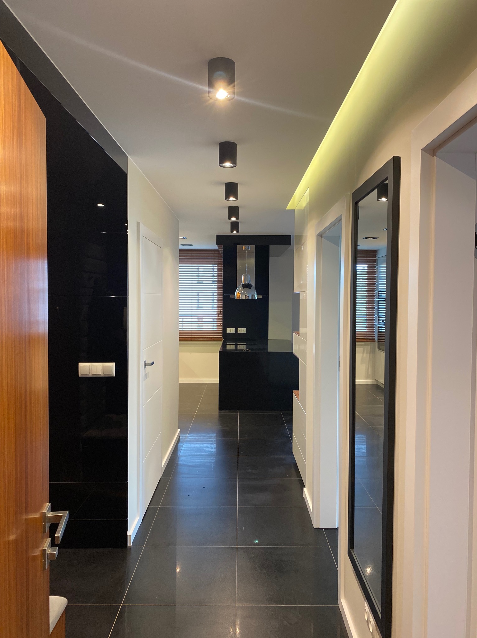 Nowoczesne wnętrze: czarne płytki podłogowe, lakierowana ściana, oświetlenie punktowe i ledowe. Widok na kuchnię z żaluzjami w tle. Minimalistyczny design.