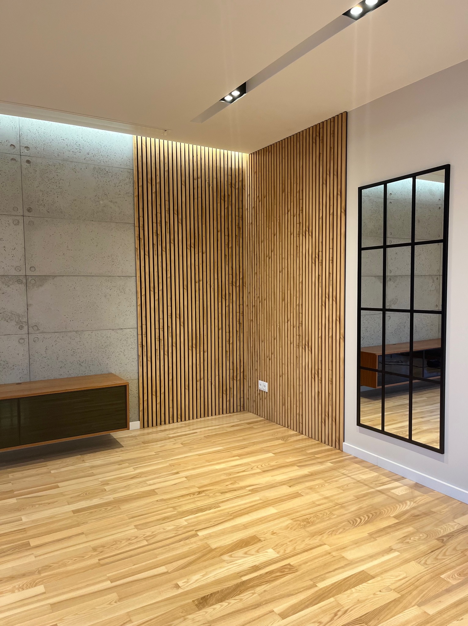 Nowoczesne wnętrze: drewniana podłoga, ściana z betonowych paneli, lamelowa boazeria i lustro w czarnej ramie. Minimalistyczny design z oświetleniem punktowym.