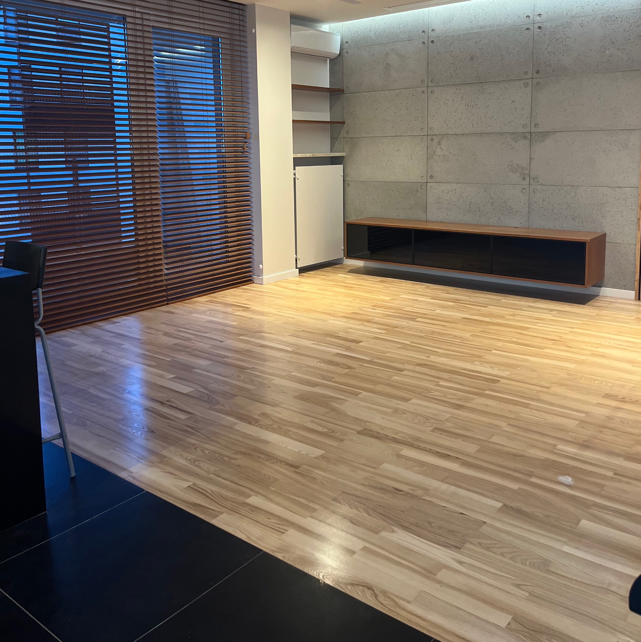 Jasne panele podłogowe ułożone w przestronnym wnętrzu z betonową ścianą i drewnianymi żaluzjami. Minimalistyczny design, nowoczesne wykończenie.