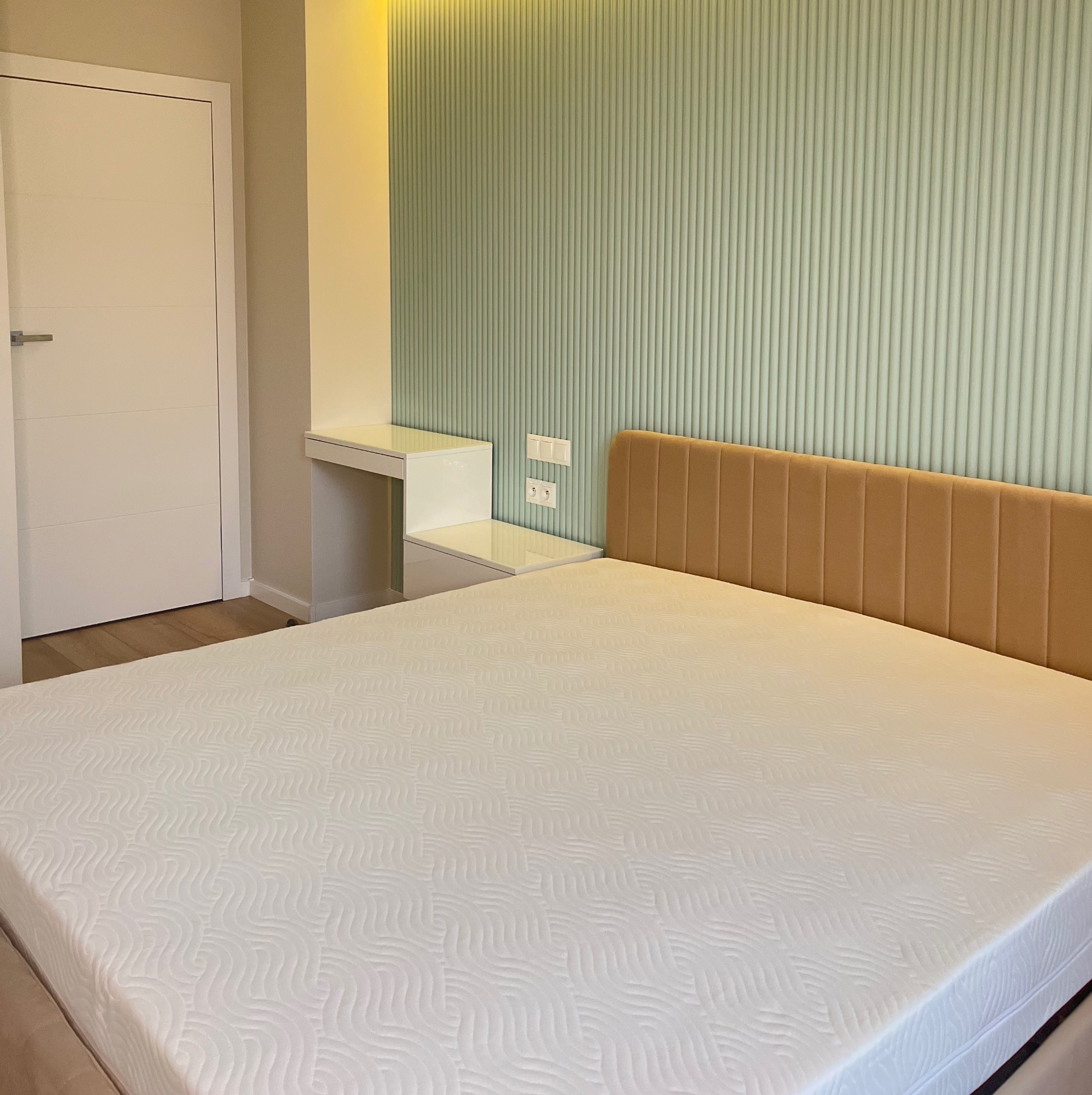 Nowoczesna sypialnia z łóżkiem z beżowym zagłówkiem, białą pościelą i ścianą w pastelowe pionowe panele. Obok biała toaletka i drzwi. Minimalistyczny design.