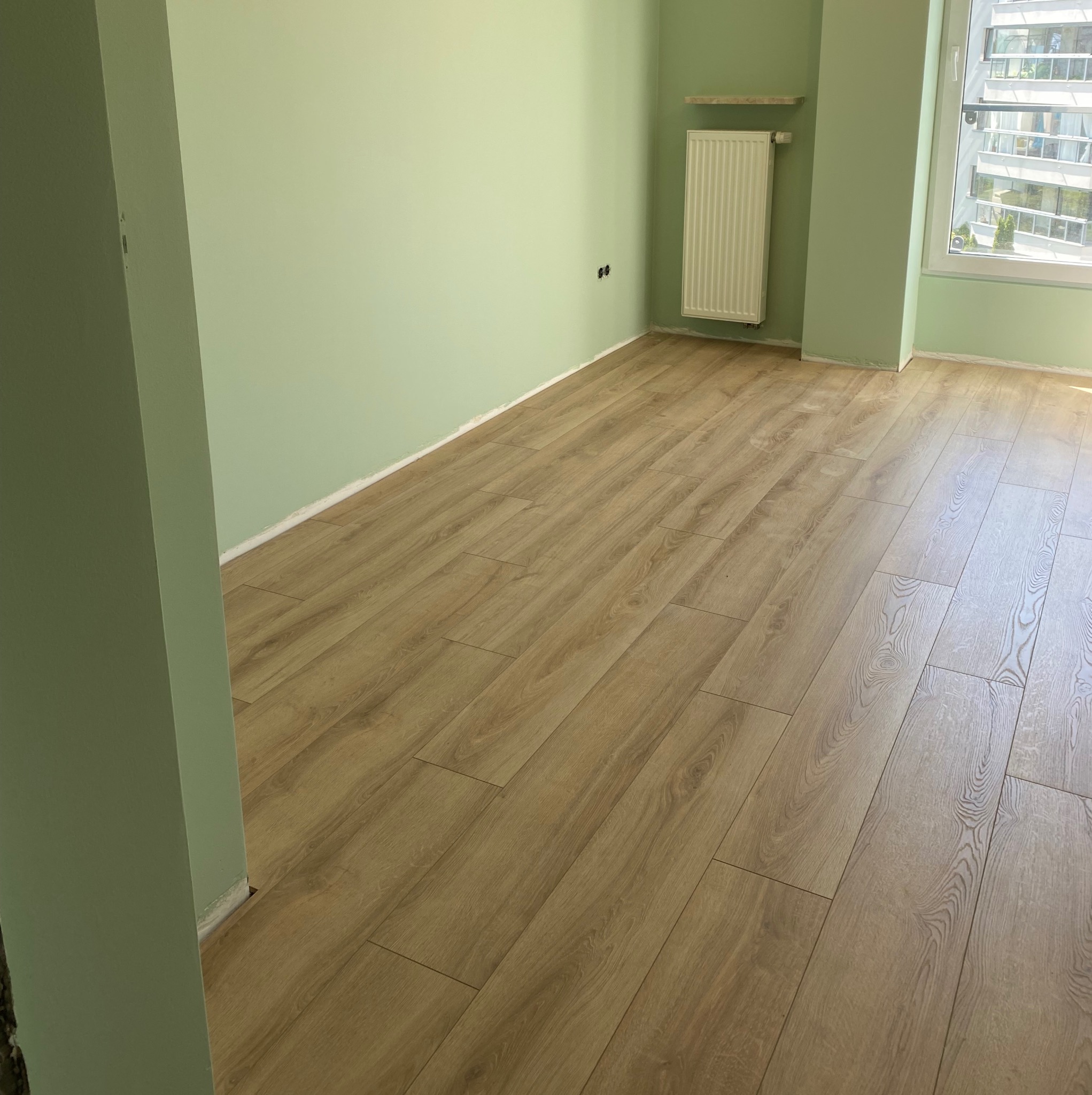 Ułożone panele podłogowe w jasnym odcieniu drewna w pokoju z zielonymi ścianami i białym grzejnikiem. Widok na okno z budynkiem w tle.