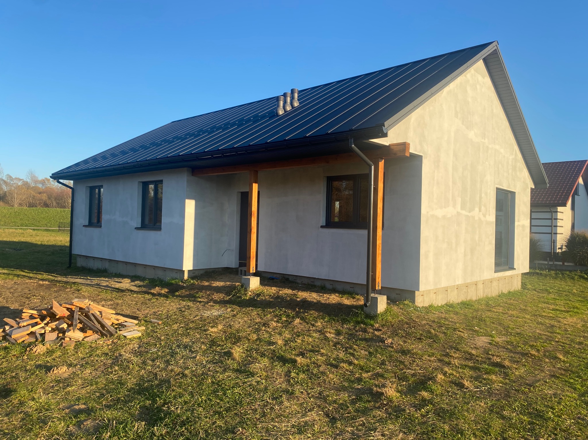 Parterowy dom w stanie surowym zamkniętym z ciemnym dachem i drewnianą konstrukcją werandy. Na trawniku leżą resztki drewna. Błękitne niebo w tle.