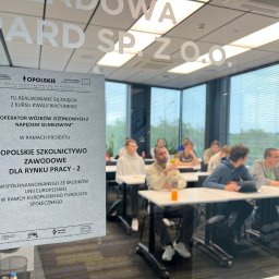 GEPARD SPÓŁKA Z OGRANICZONĄ ODPOWIEDZIALNOŚCIĄ - Sala szkoleniowa z uczestnikami kursu 'Operator wózków jezdniowych', widoczna przez szybę z naklejonym plakatem informacyjnym o projekcie 'Opolskie Szkolnictwo Zawodowe'.