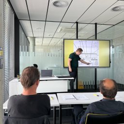 GEPARD SPÓŁKA Z OGRANICZONĄ ODPOWIEDZIALNOŚCIĄ - Sala szkoleniowa: instruktor omawia schemat wózka widłowego na ekranie, uczestnicy siedzą przy stołach z notatkami. Nowoczesne wnętrze, jasne oświetlenie.