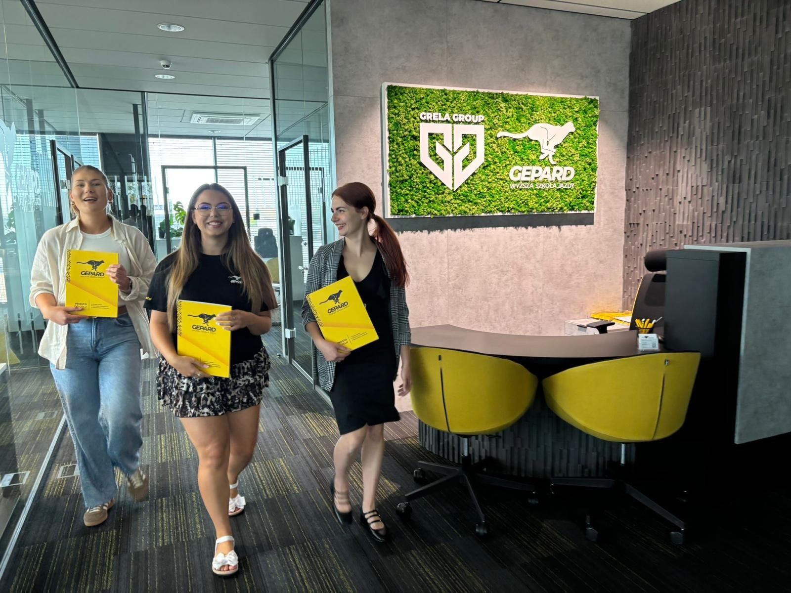 Trzy uśmiechnięte kobiety z żółtymi teczkami z logo Gepard idą korytarzem biura z recepcją i logo firmy na ścianie. Jasne wnętrze, nowoczesny design.