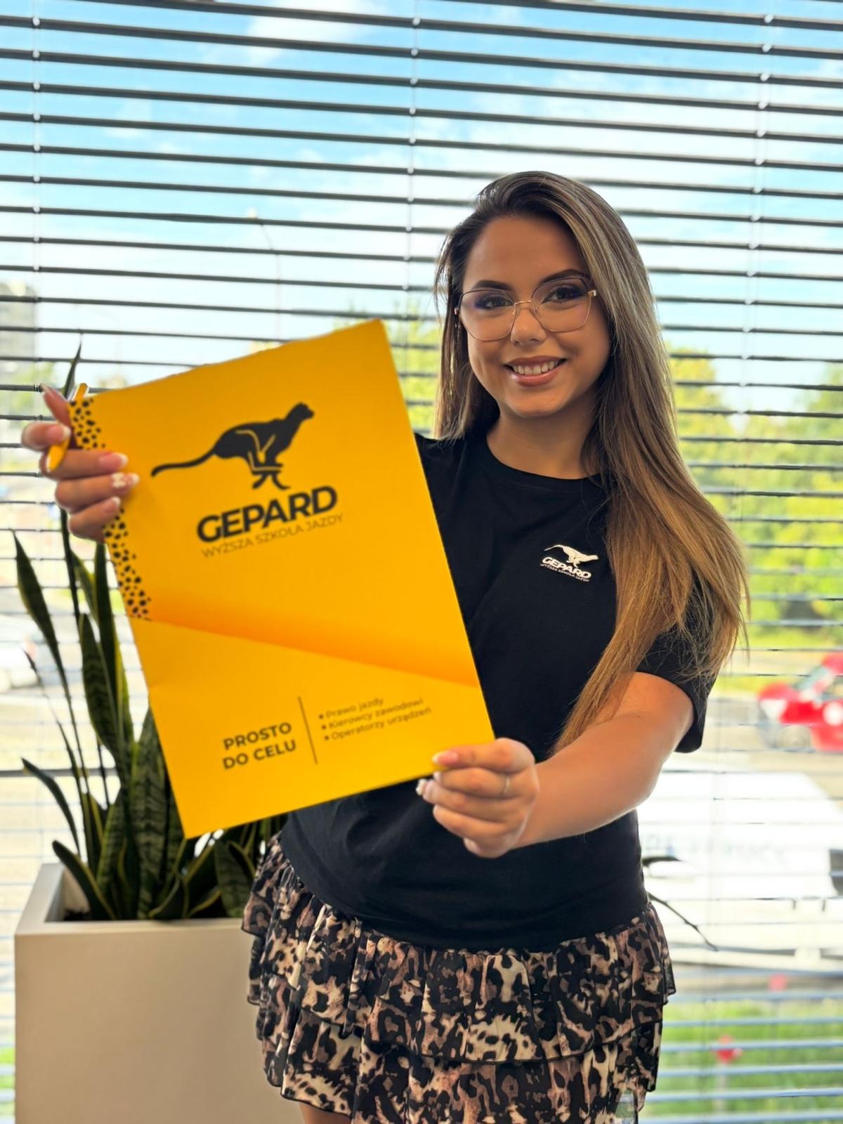 Uśmiechnięta kobieta w okularach prezentuje żółty folder z logo szkoły jazdy Gepard, na tle biura z żaluzjami i rośliną doniczkową.