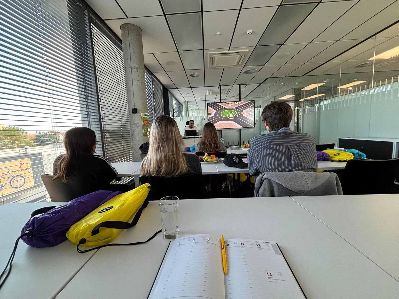 Sala szkoleniowa z uczestnikami kursu, widok zza stołu z notatnikiem i długopisem. Na ekranie prezentacja skrzyżowania drogowego, nowoczesne wnętrze z dużymi oknami.