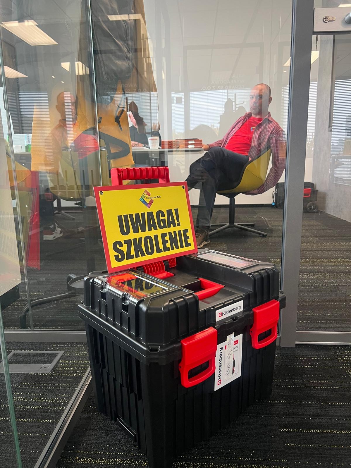 Czarna skrzynka narzędziowa Kistenberg z żółtą tabliczką 'Uwaga! Szkolenie' w biurze, w tle siedzący mężczyzna na krześle. Widok przez szklaną ścianę.