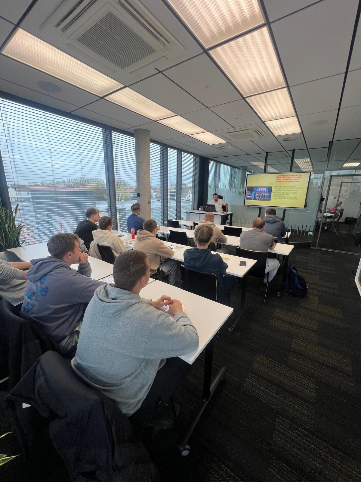 Sala szkoleniowa z uczestnikami siedzącymi przy biurkach, prowadzący prezentuje materiał na ekranie. Widok z perspektywy uczestnika.
