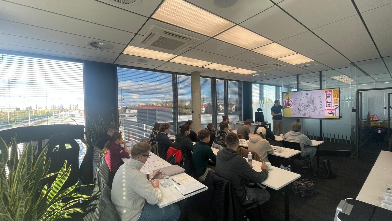 Sala szkoleniowa z widokiem na miasto. Instruktor prezentuje schemat wózka widłowego na ekranie, a kursanci siedzą przy biurkach i notują. Nowoczesne wnętrze z dużymi oknami.