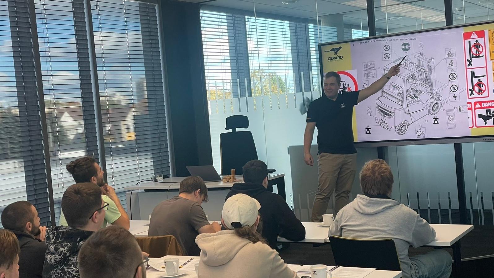 Szkolenie z obsługi wózków widłowych: instruktor prezentuje schemat na interaktywnej tablicy, uczestnicy siedzą przy stołach z notatkami i filiżankami.