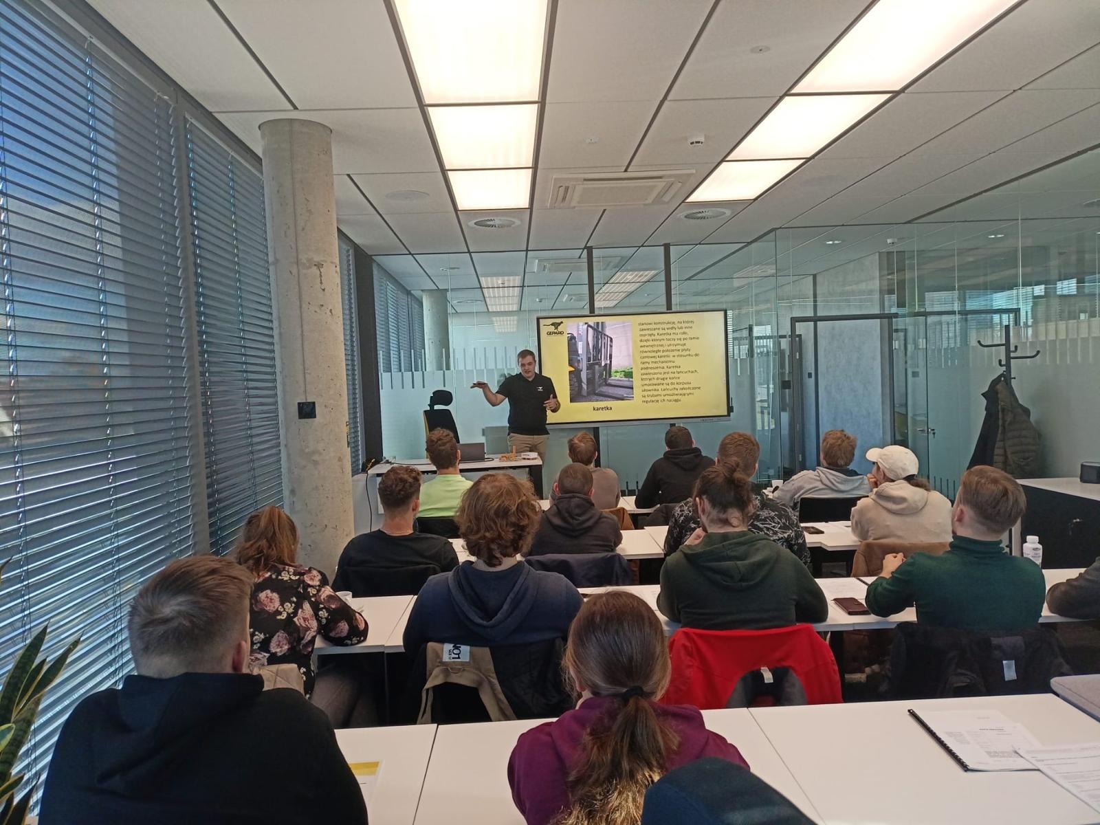 Sala szkoleniowa z widokiem na prezentację o wózkach widłowych. Instruktor gestykuluje, a uczestnicy siedzą przy stołach. Nowoczesne wnętrze z żaluzjami.