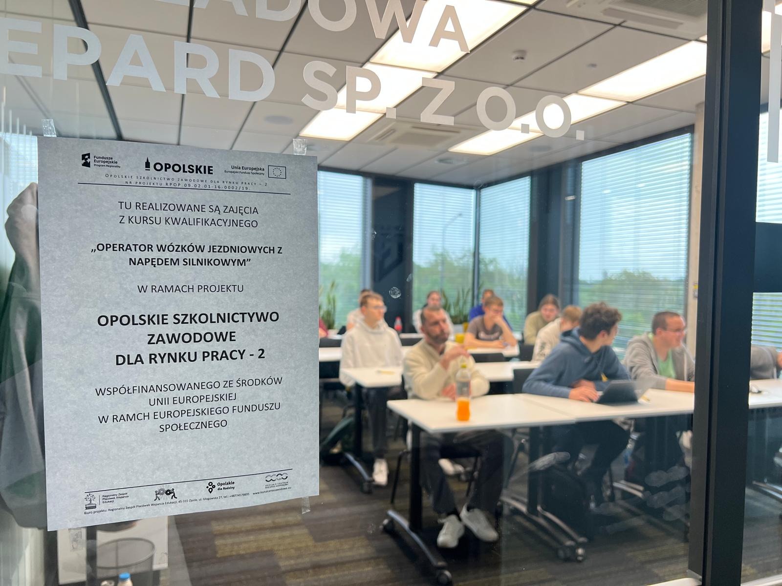 Sala szkoleniowa z uczestnikami kursu 'Operator wózków jezdniowych', widoczna przez szybę z naklejonym plakatem informacyjnym o projekcie 'Opolskie Szkolnictwo Zawodowe'.