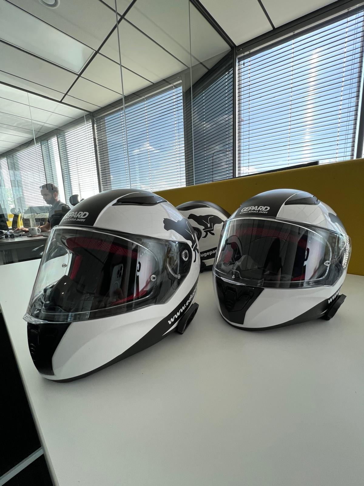 Trzy biało-czarne kaski motocyklowe z logo geparda, ustawione na stole w jasnym pomieszczeniu z widokiem na miasto przez żaluzje. Widoczny pracownik w tle.