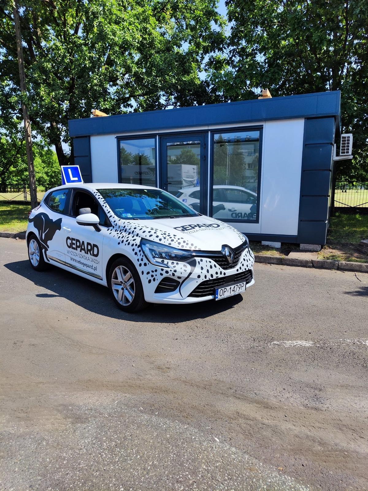 Biały samochód z oznaczeniem 'L' i logo szkoły jazdy 'Gepard' na tle biura. Auto z czarnymi plamkami, imitującymi geparda, zaparkowane na asfalcie w słoneczny dzień.