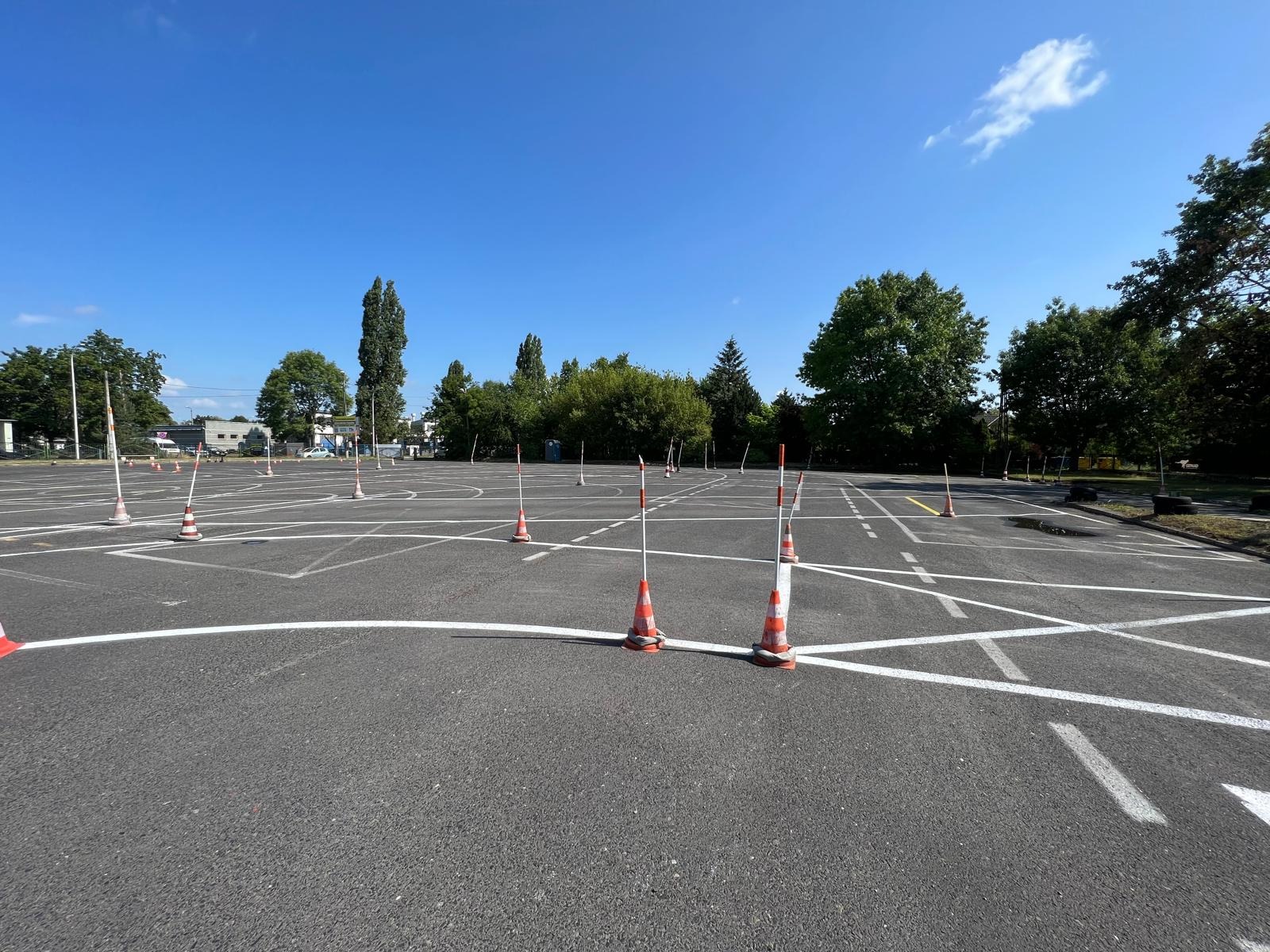 Plac manewrowy z białymi liniami i pomarańczowo-białymi pachołkami, przygotowany do szkolenia kierowców w słoneczny dzień. Widok na drzewa i błękitne niebo.