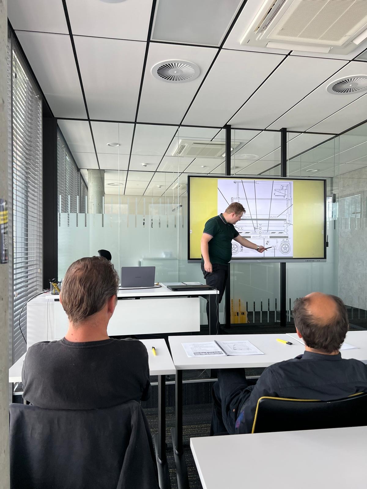 Sala szkoleniowa: instruktor omawia schemat wózka widłowego na ekranie, uczestnicy siedzą przy stołach z notatkami. Nowoczesne wnętrze, jasne oświetlenie.