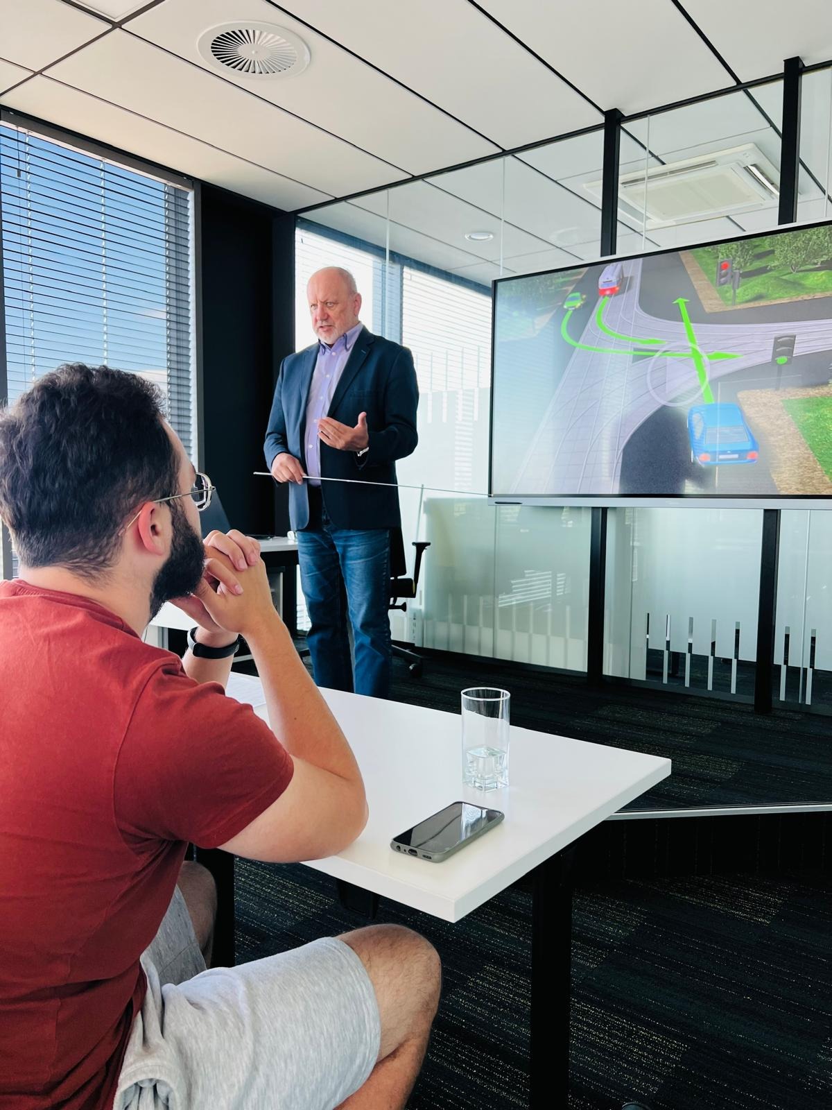 Prelegent w marynarce prezentuje symulację drogową na ekranie podczas kursu. Uczestnik w czerwonej koszulce słucha. Nowoczesne biuro z dużymi oknami.