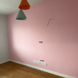 Ares Bud Hennadii Deleraikov - Różowa ściana z gniazdkami i wystającymi kablami, drewniana podłoga, biała listwa przypodłogowa, wiszące lampy w kształcie stożków w kolorach pomarańczowym i miętowym.