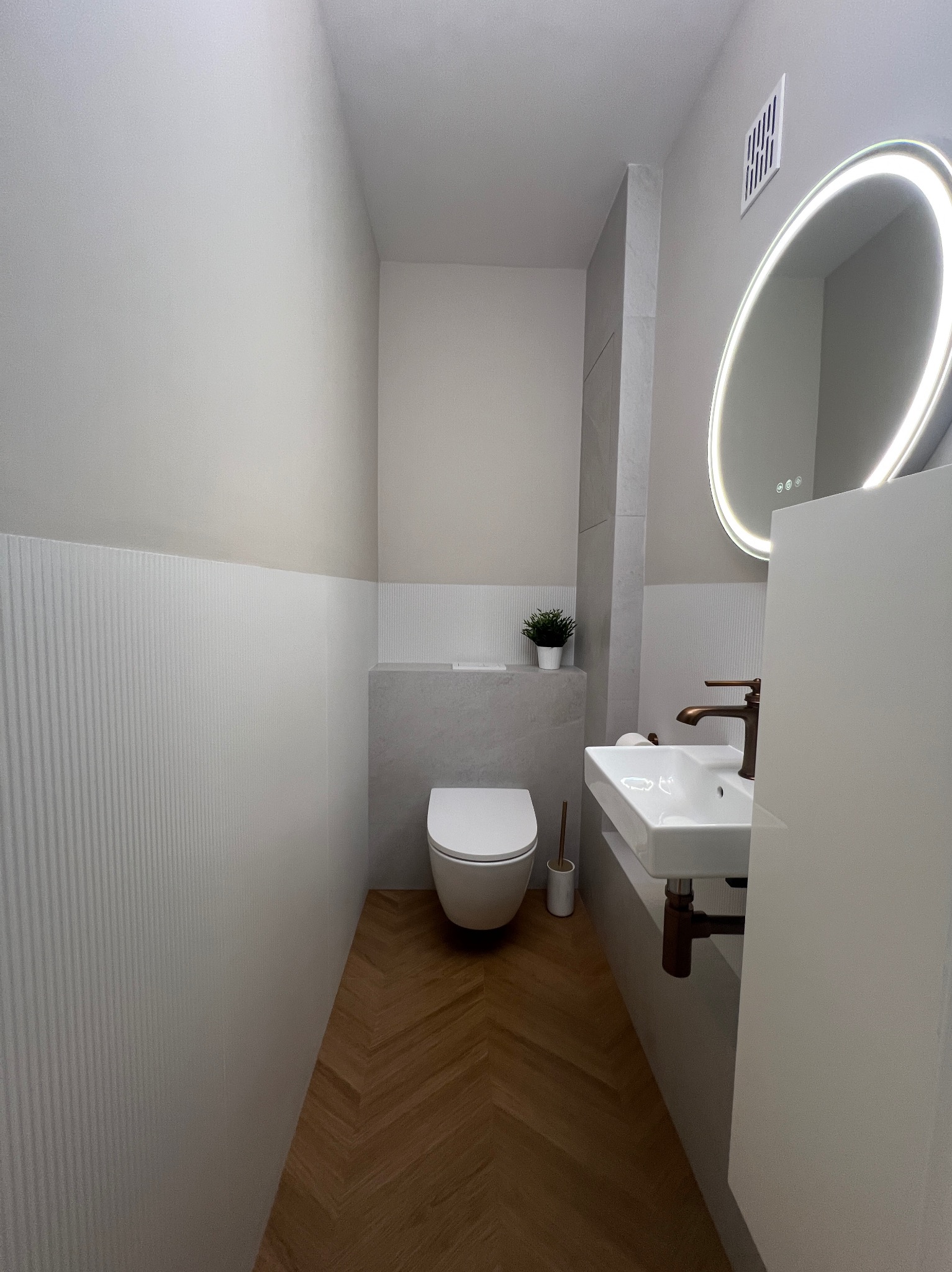 Nowoczesna toaleta: biała armatura, drewniana podłoga w jodełkę, lustro z podświetleniem LED i minimalistyczne detale. Ściany wykończone w odcieniach szarości.