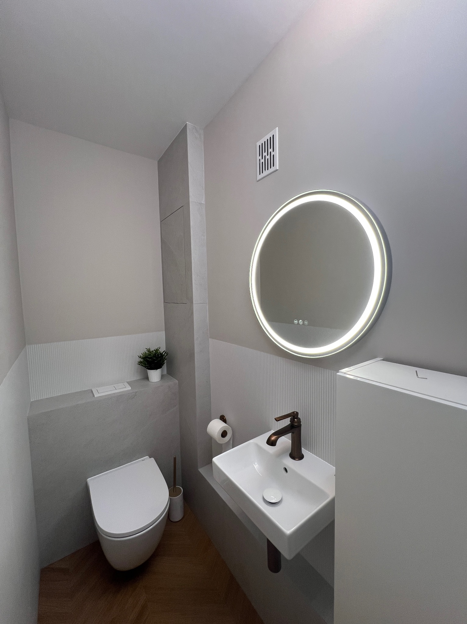 Nowoczesna łazienka w szarościach z drewnianą podłogą. Uwagę przykuwa okrągłe lustro z podświetleniem LED i minimalistyczna umywalka z brązową armaturą.