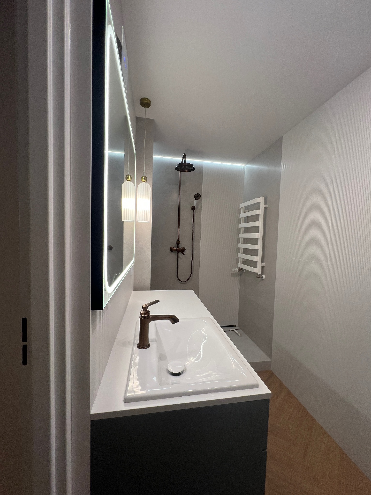 Nowoczesna łazienka z podświetlanym lustrem, brązową armaturą i grzejnikiem drabinkowym. Minimalistyczny design z drewnianą podłogą i subtelnym oświetleniem LED.