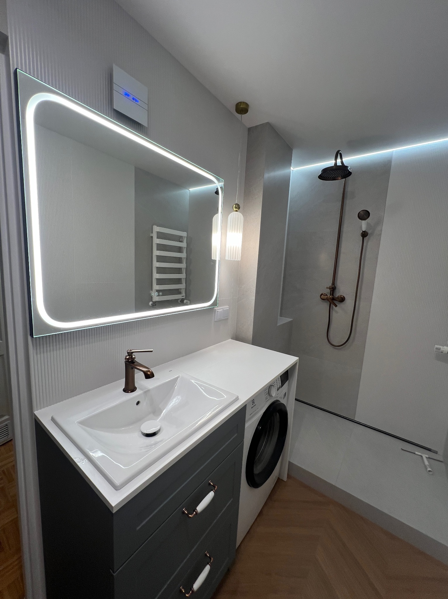 Nowoczesna łazienka po remoncie: szara szafka z umywalką, podświetlane lustro, pralka i prysznic z deszczownicą w odcieniach szarości i brązu. Parkiet w jodełkę.