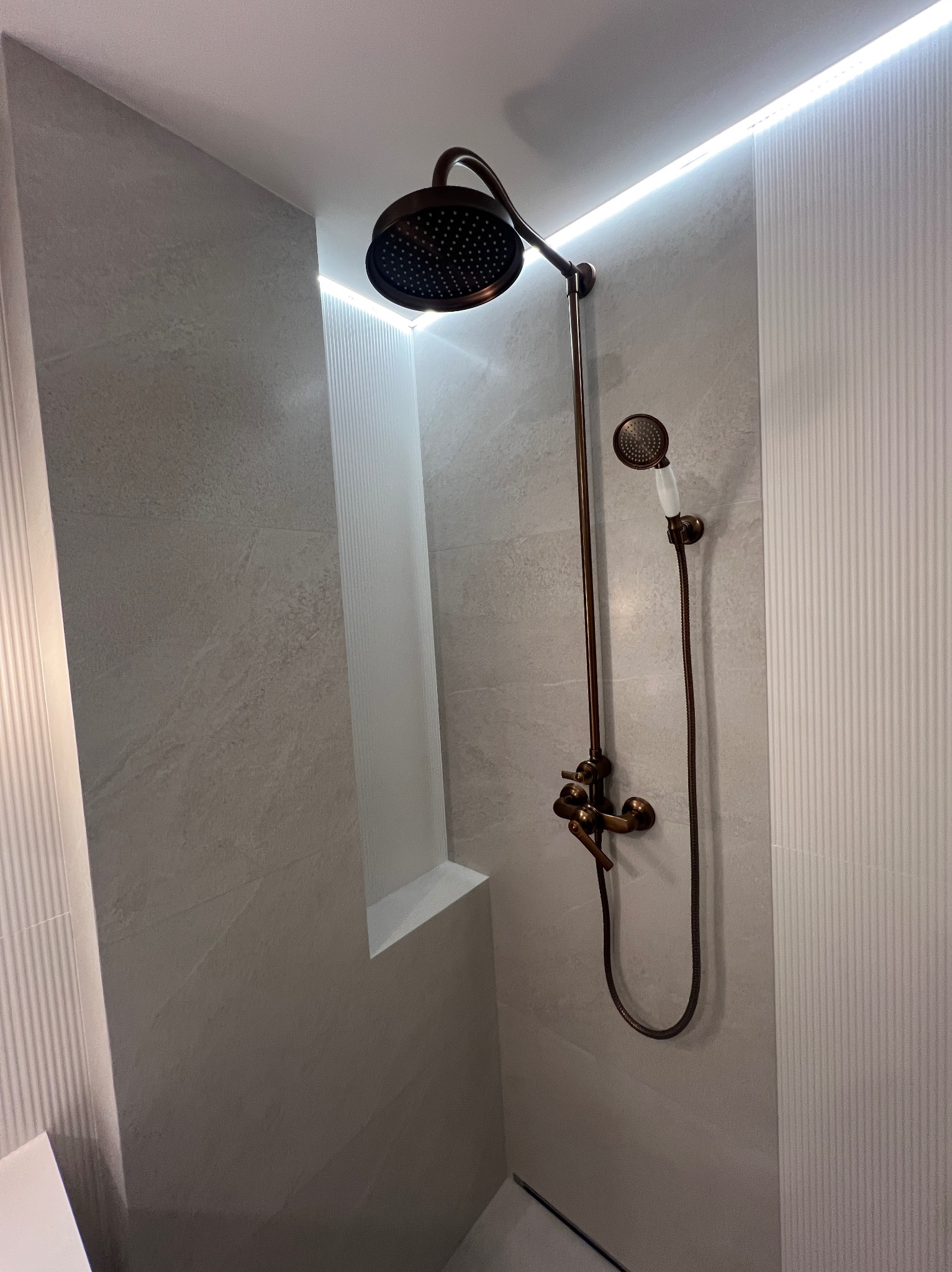 Wykończona łazienka z szarymi płytkami i miedzianą armaturą prysznicową. Podświetlane wnęki i pionowe panele dekoracyjne. Nowoczesny design.