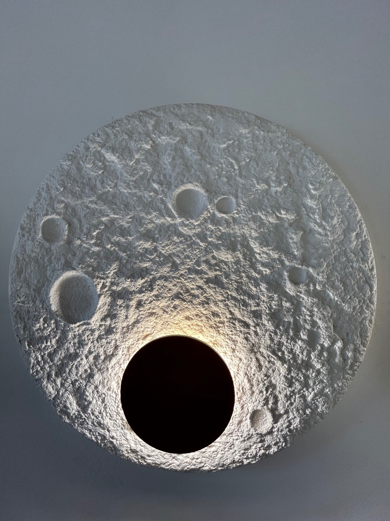 Dekoracyjna, okrągła lampa ścienna z fakturą przypominającą powierzchnię księżyca, z centralnym otworem emitującym ciepłe światło. Minimalistyczny design.