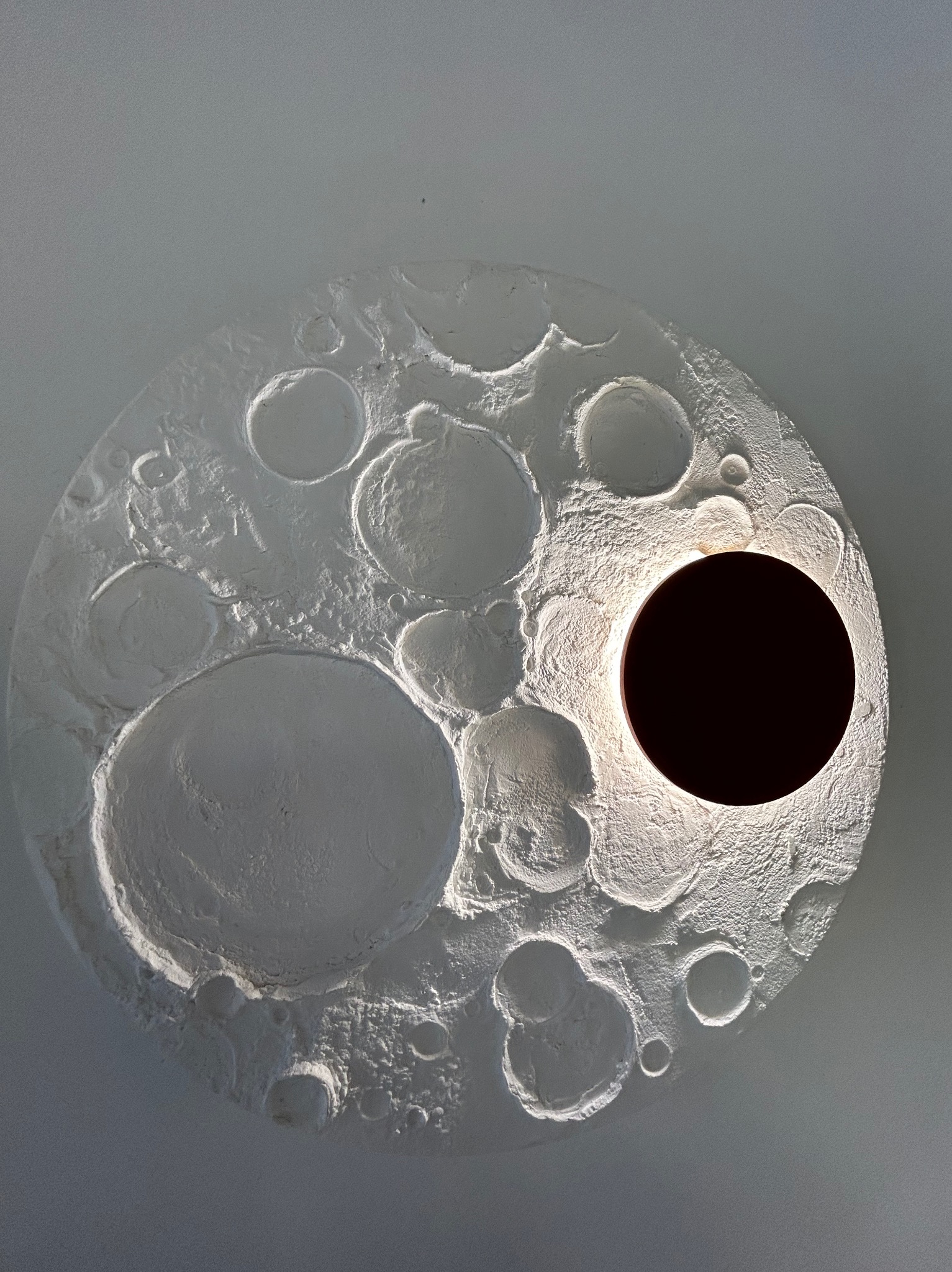 Dekoracyjna lampa sufitowa w kształcie księżyca z kraterami, oświetlona od wewnątrz, tworząca unikalny efekt wizualny. Biała, teksturowana powierzchnia.