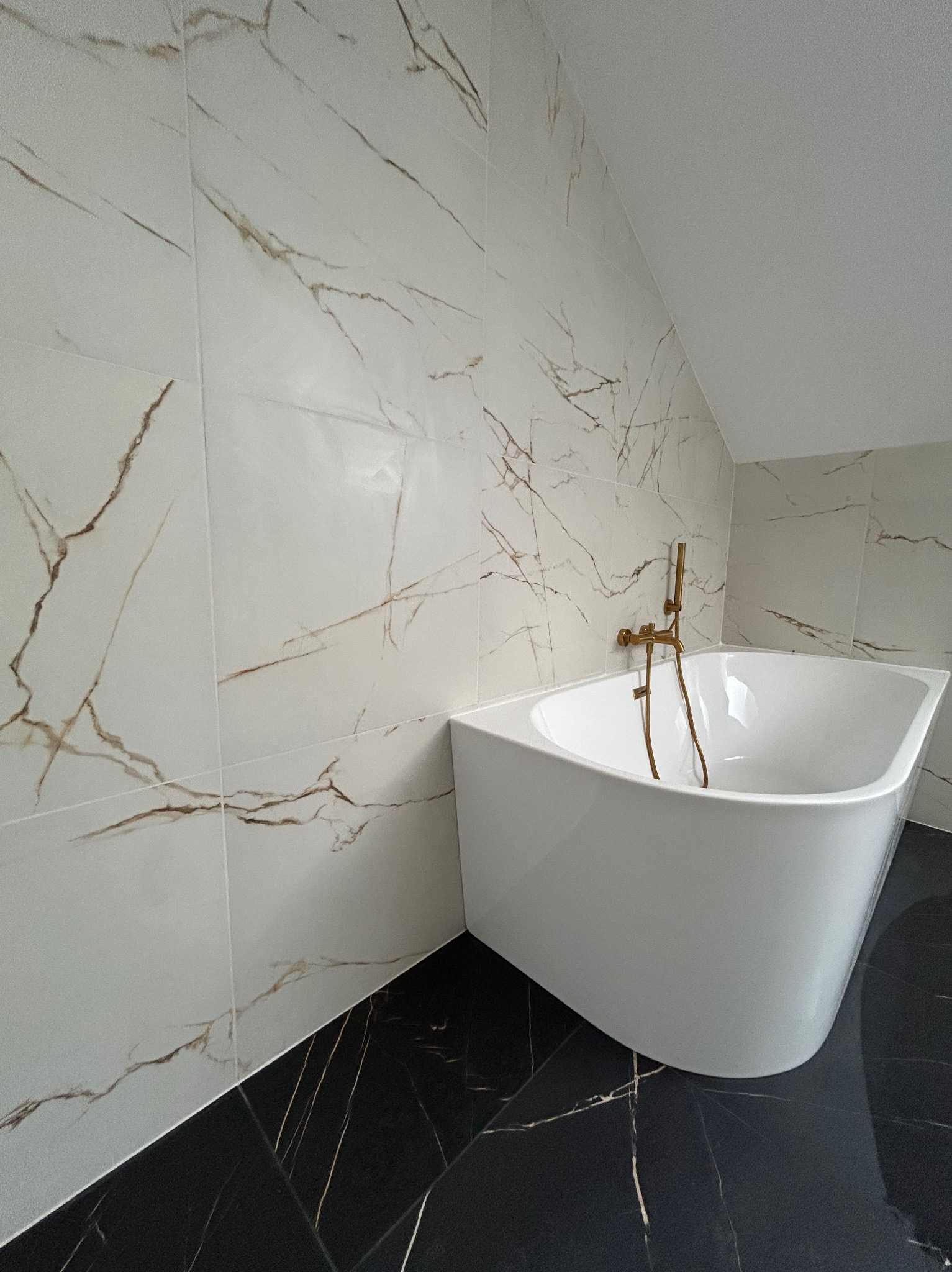 Biała wanna narożna z złotą armaturą na tle ściany i podłogi z płytek imitujących marmur w łazience na poddaszu.