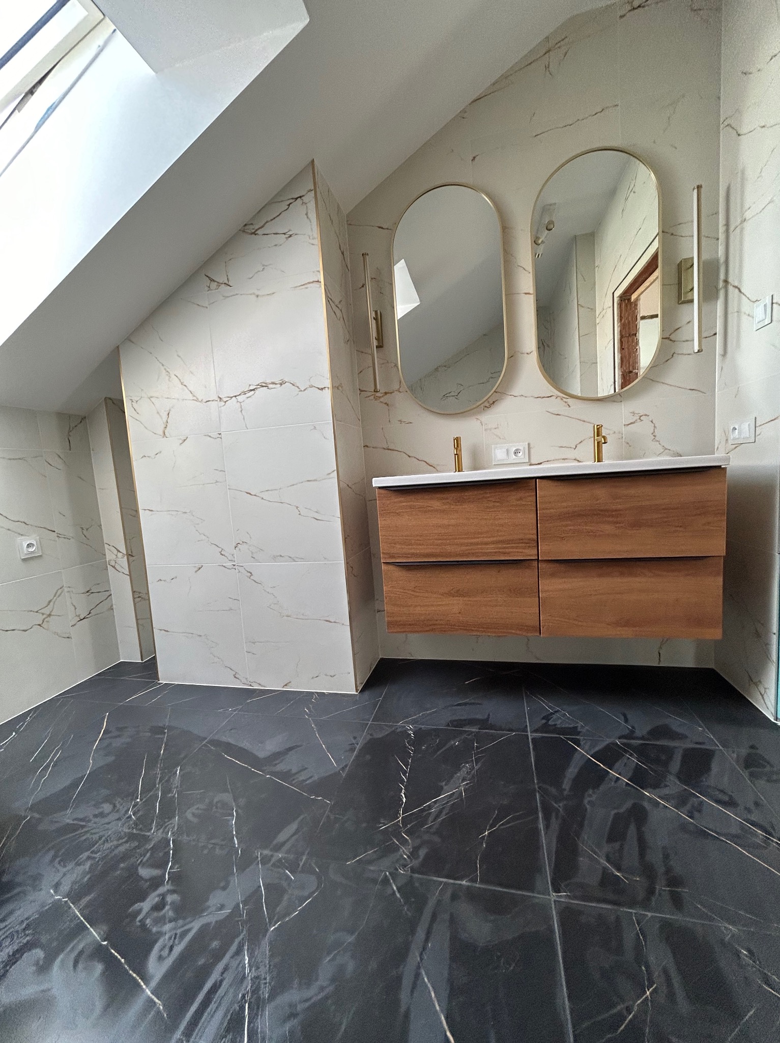 Wykończona łazienka na poddaszu z czarną marmurową podłogą i białymi płytkami ściennymi z żyłkami. Dwie owalne lustra nad drewnianą szafką z umywalkami.