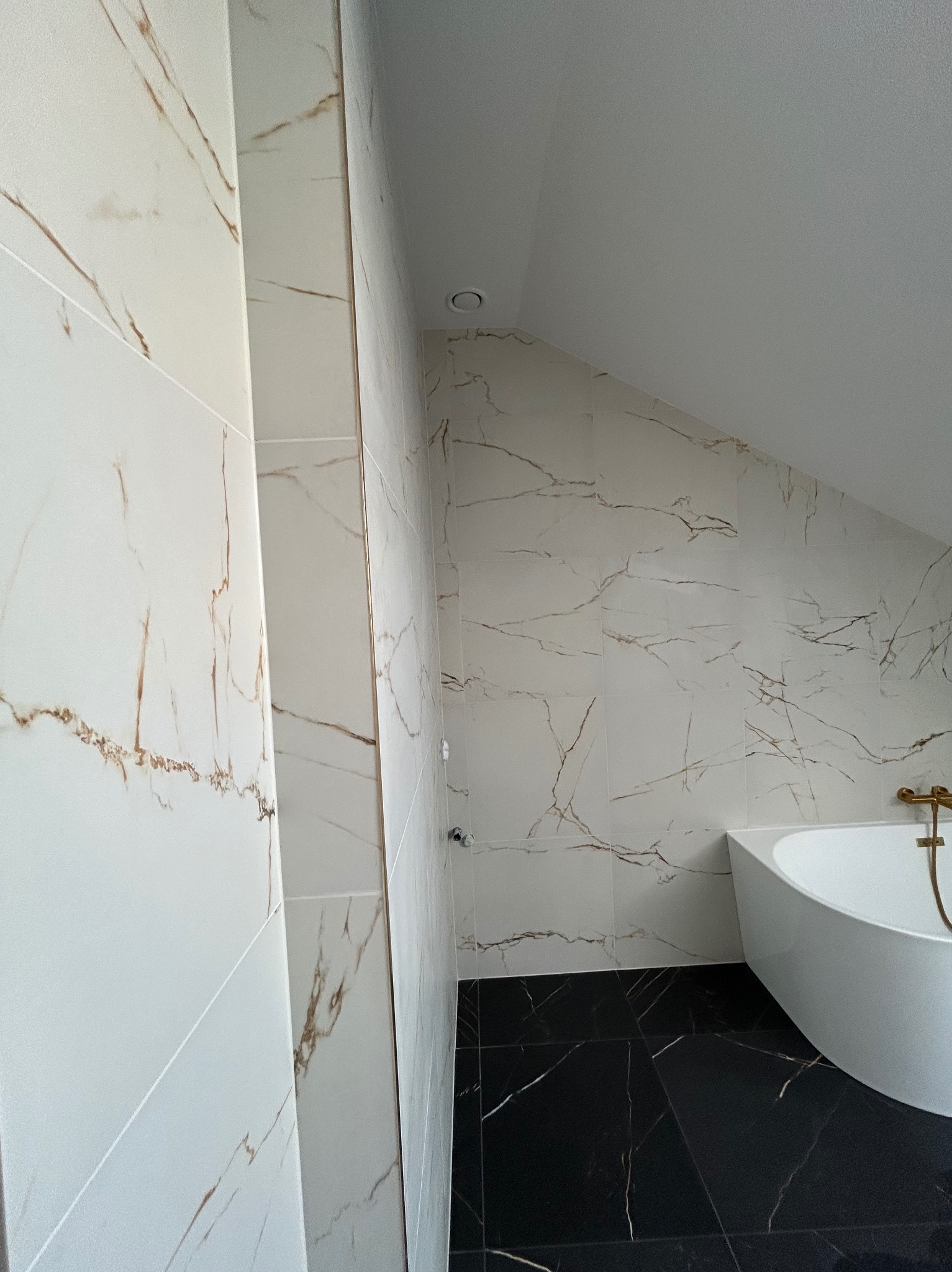 Wykończona łazienka na poddaszu z białymi płytkami imitującymi marmur z brązowymi żyłami na ścianach i czarną podłogą z białymi żyłami. Widoczna wanna.