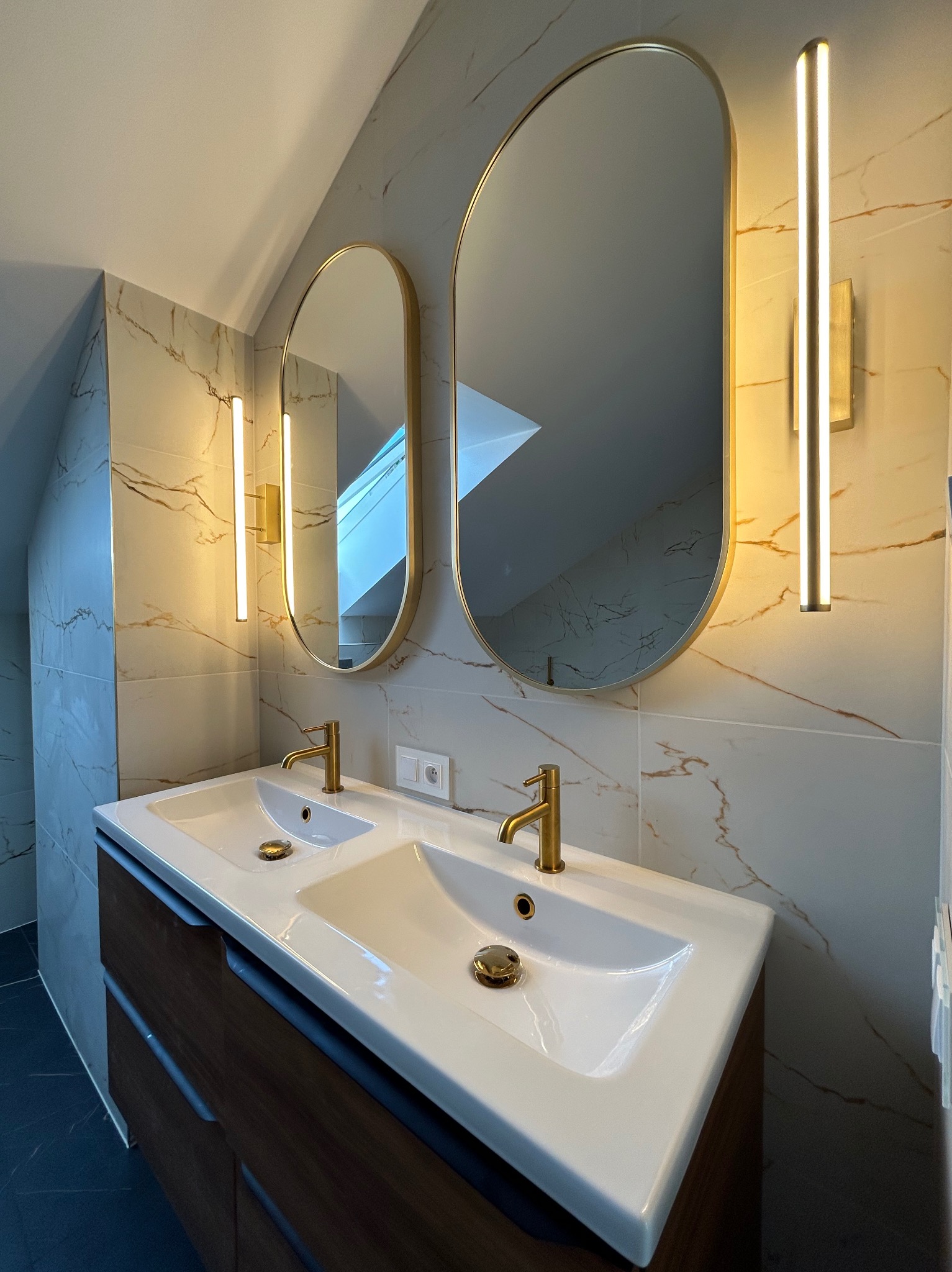 Nowoczesna łazienka na poddaszu: marmurowe płytki, owalne lustra w złotych ramach, podwójna umywalka z szafką w kolorze drewna i złote baterie.