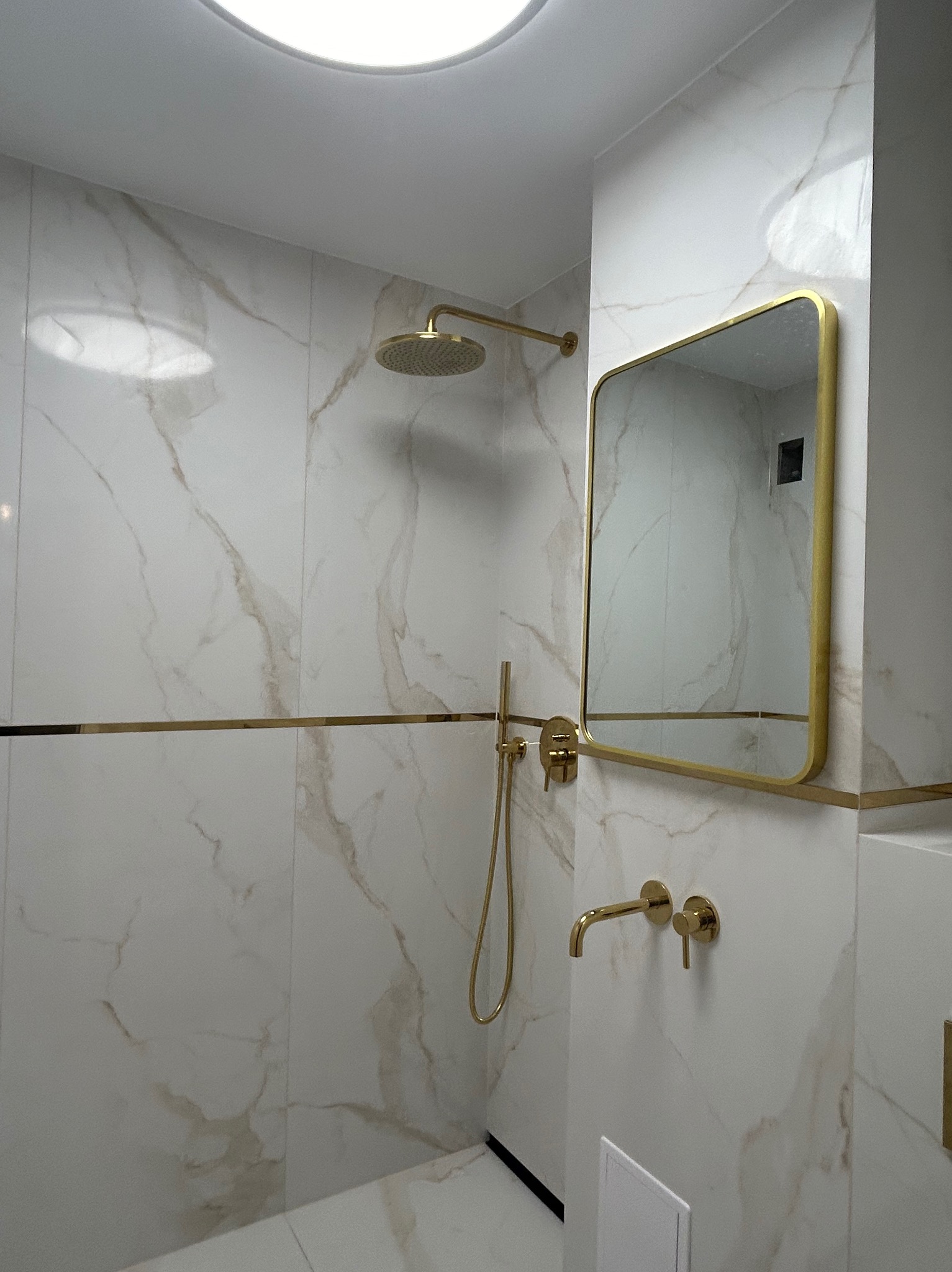 Elegancka łazienka z marmurowymi płytkami, złotą armaturą i lustrem w złotej ramie. Minimalistyczny design, nowoczesne oświetlenie sufitowe.