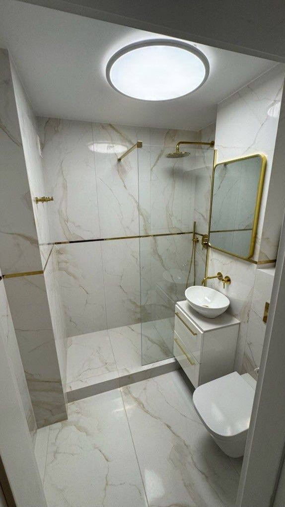 Elegancka łazienka z kabiną prysznicową typu walk-in, wykończona białym marmurem z złotymi akcentami. Umywalka nablatowa i lustro w złotej ramie dodają luksusowego charakteru.