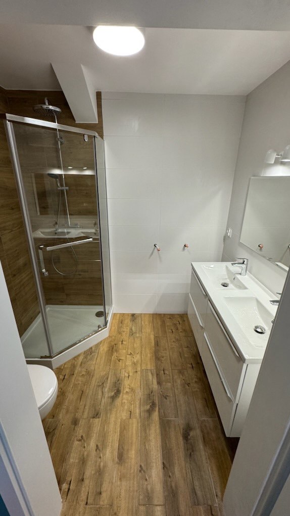Nowoczesna łazienka po remoncie: kabina prysznicowa z drewnopodobnymi płytkami, białe płytki ścienne, podwójna umywalka z szafką i drewniana podłoga.