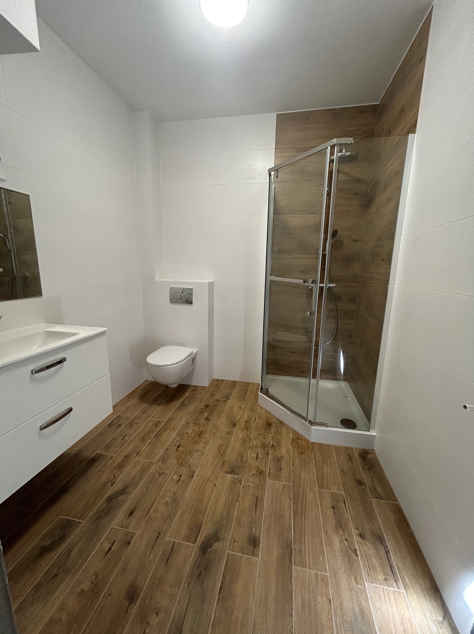 Nowoczesna łazienka z kabiną prysznicową, umywalką z szafką i podwieszaną toaletą. Brązowe płytki imitujące drewno na podłodze i częściowo na ścianie kabiny.