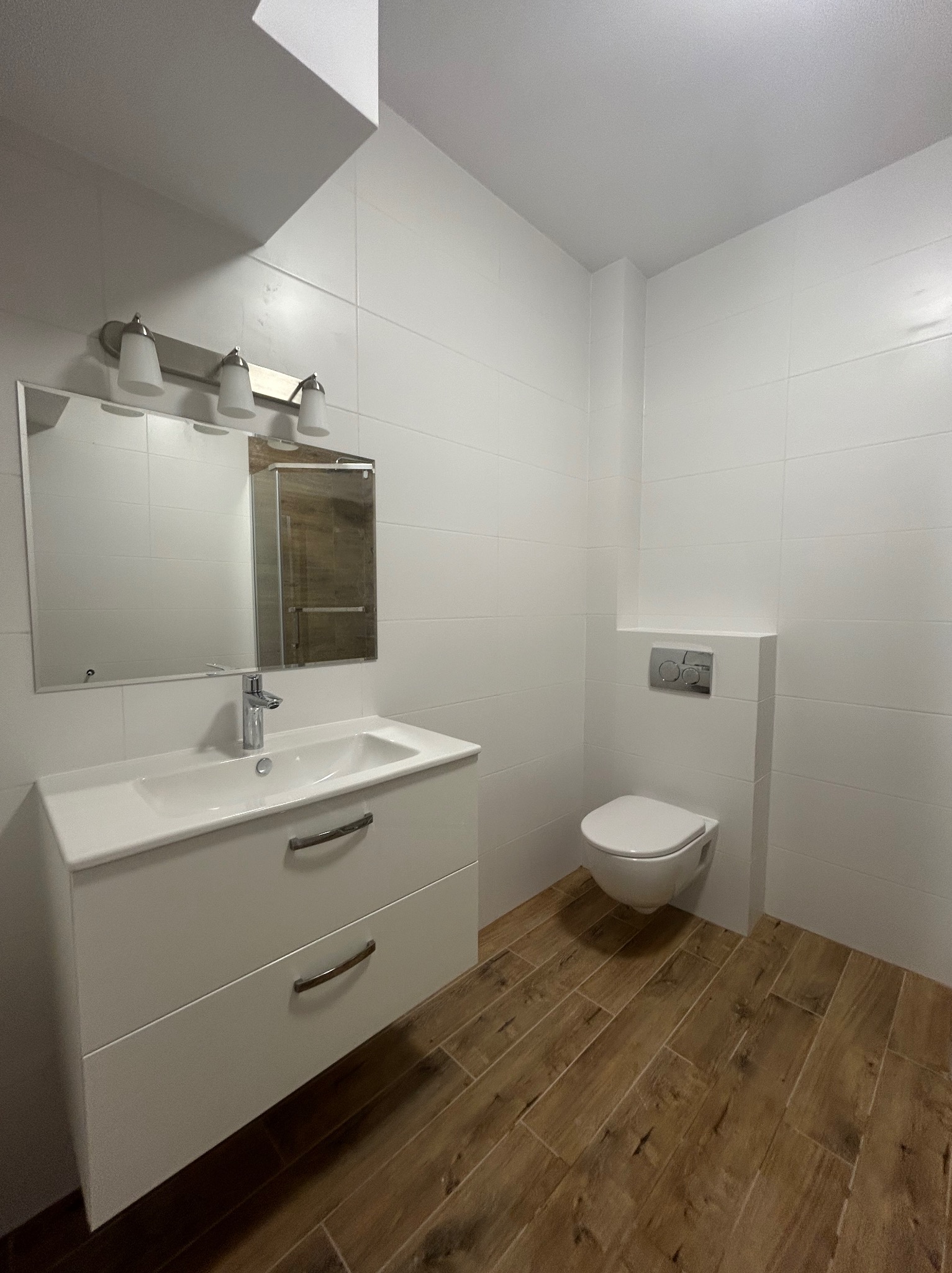 Nowoczesna łazienka z białymi płytkami i drewnopodobną podłogą. Umywalka z szafką, lustro z oświetleniem i wisząca toaleta tworzą spójną całość.