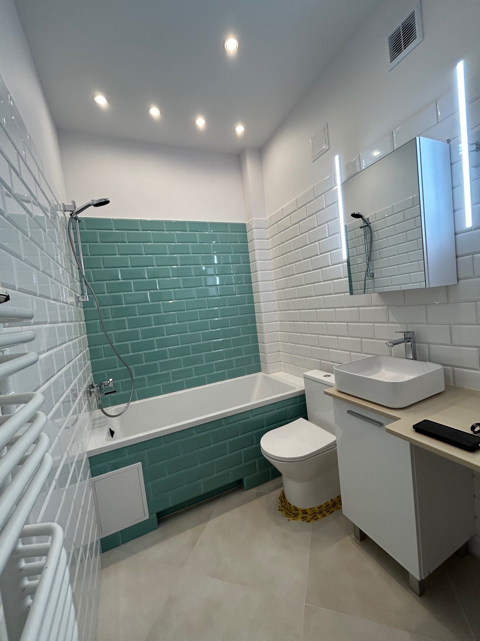 Wykończona łazienka z wanną obłożoną turkusową glazurą typu metro, białą umywalką nablatową i grzejnikiem drabinkowym. Nowoczesne oświetlenie punktowe i lustro z podświetleniem LED.