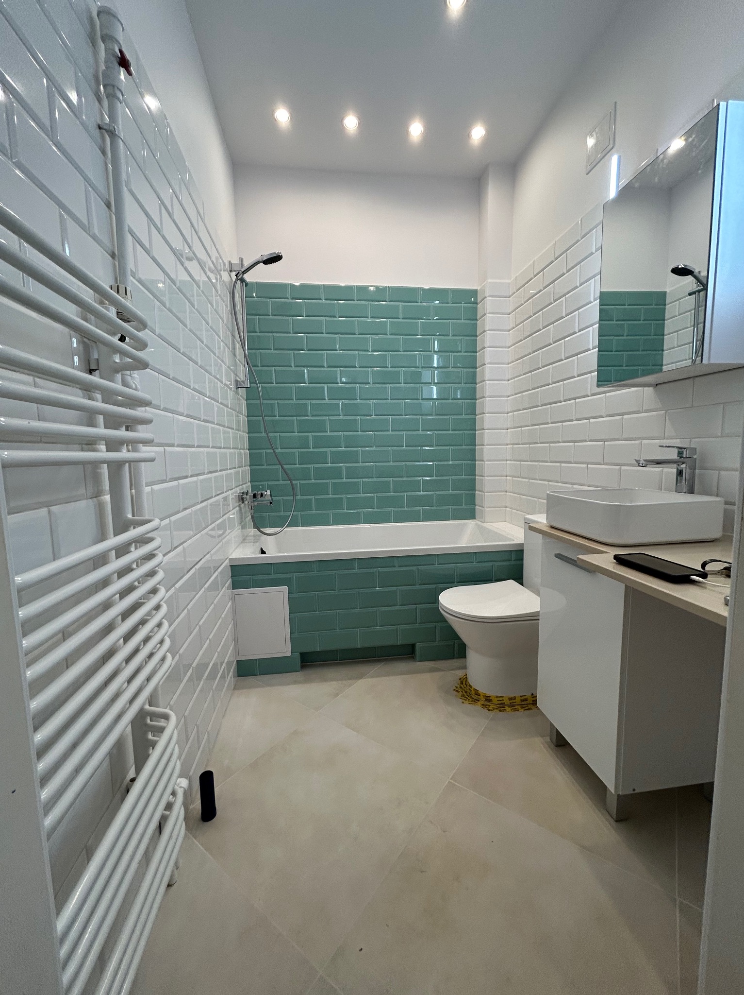 Wykończona łazienka z biało-turkusowymi płytkami typu metro, grzejnikiem drabinkowym i białymi meblami łazienkowymi. Widoczne detale wykończeniowe.