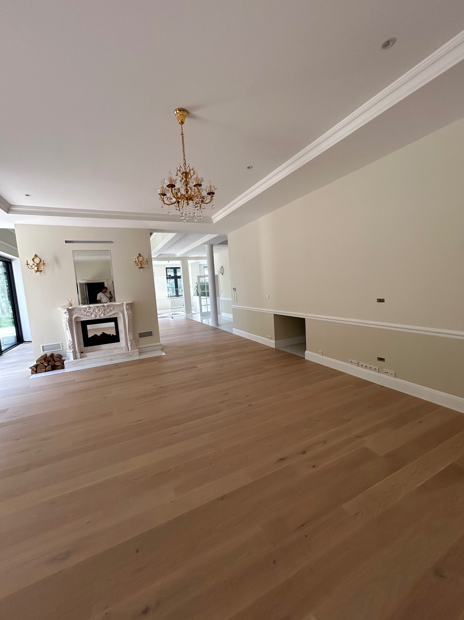 Jasne, przestronne wnętrze z drewnianą podłogą, kremowymi ścianami z listwami, ozdobnym kominkiem i kryształowym żyrandolem. Widok na dalsze pomieszczenia.