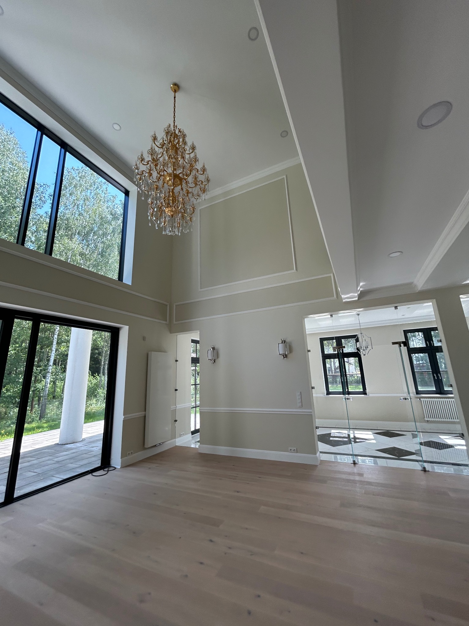 Przestronne wnętrze z wysokimi oknami i parkietem. Kryształowy żyrandol, dekoracyjne listwy ścienne i widok na zieleń za oknem. Nowoczesny design z elementami klasycznymi.