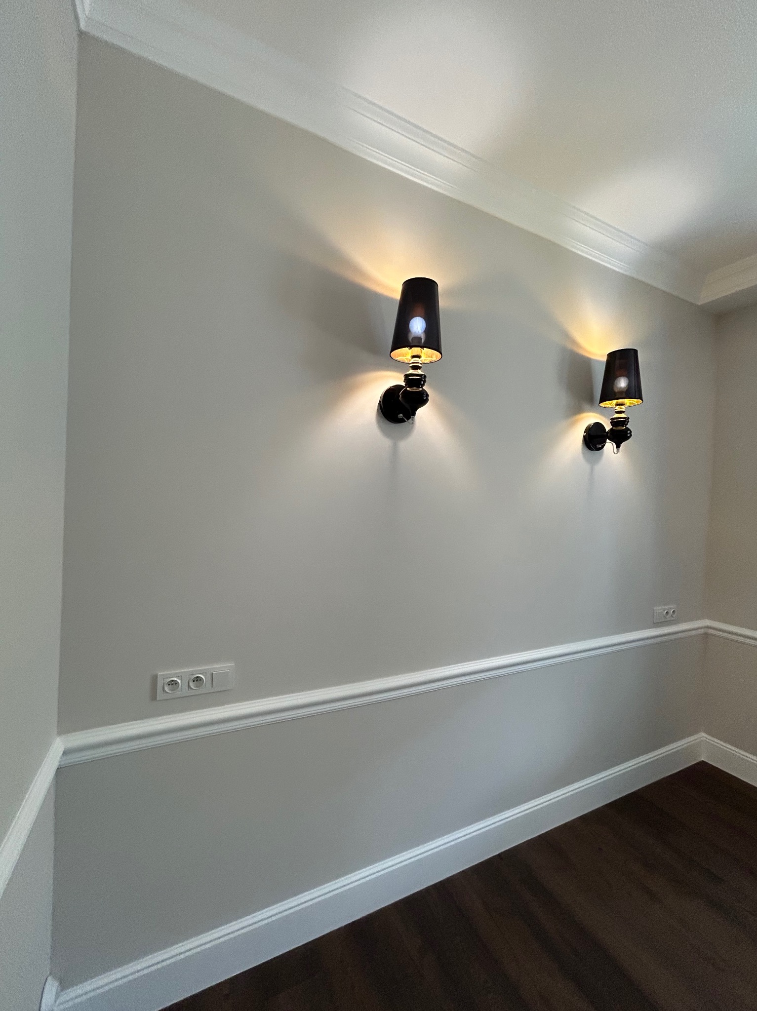 Eleganckie wnętrze z szarymi ścianami, białymi listwami przypodłogowymi i sufitowymi oraz dwoma czarnymi kinkietami, oświetlającymi przestrzeń ciepłym światłem.