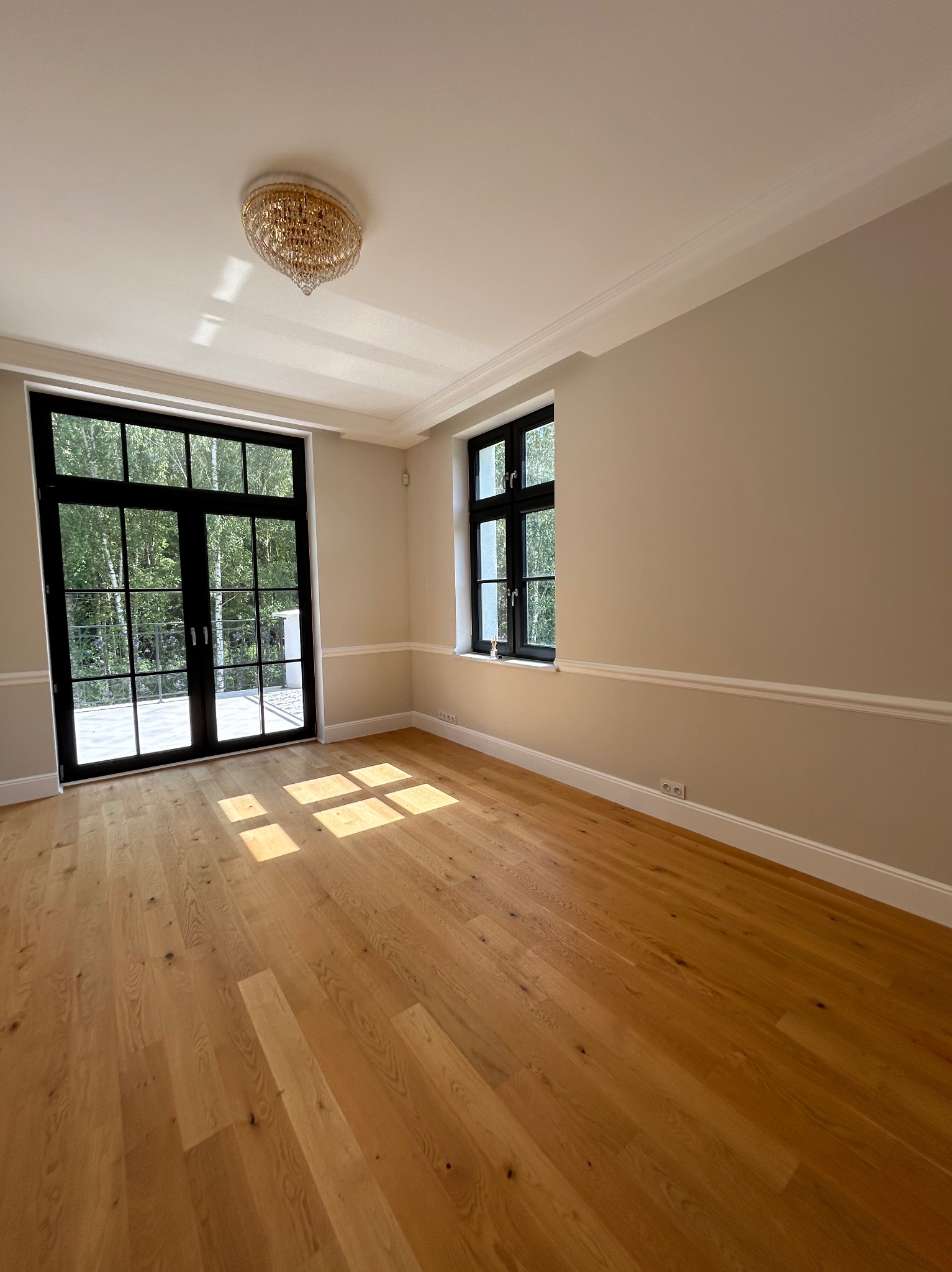 Jasne, przestronne wnętrze po remoncie: drewniana podłoga, świeżo pomalowane ściany w odcieniach beżu, czarne okna i drzwi balkonowe z widokiem na zieleń.