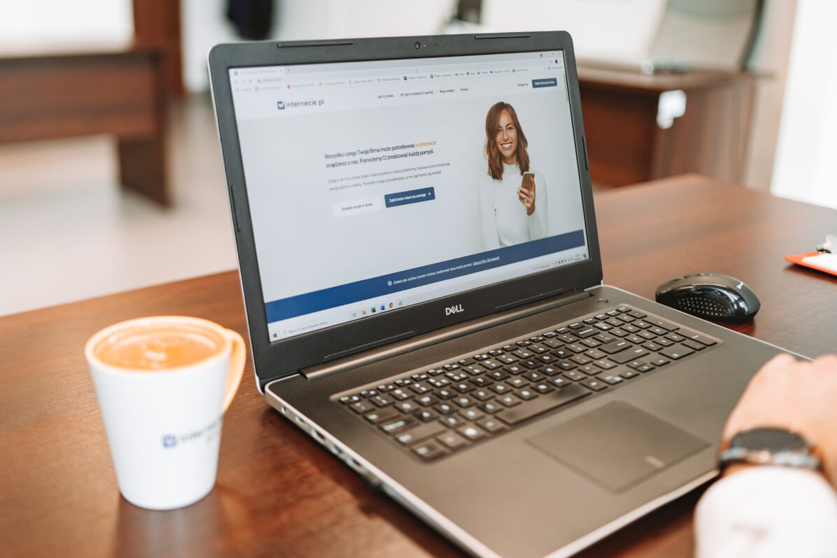 Laptop Dell wyświetla stronę internetową internecie.pl na biurku z kawą i myszką. Widoczny fragment ręki z zegarkiem. Ciepłe oświetlenie biurowe.
