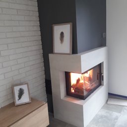 PROGRES Piotr Kuczyński Usługi Ogólnobudowlane - Nowoczesny kominek z paleniskiem w salonie w Zdunach. Biała cegła na ścianie, minimalistyczny design, płomienie widoczne przez szybę. Element wykończenia wnętrza.