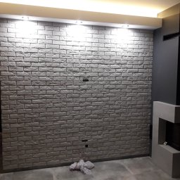 PROGRES Piotr Kuczyński Usługi Ogólnobudowlane - Ściana z dekoracyjnej cegły w szarościach, oświetlona punktowo, z fragmentem nowoczesnego kominka po prawej stronie. Widoczne gniazdka elektryczne i fragment podłogi.